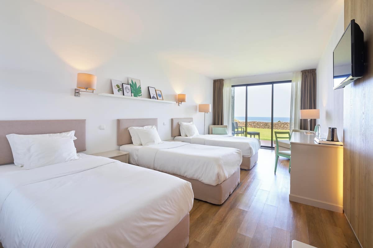 A hotel room with three beds, a TV, a desk, and a large window with a view.