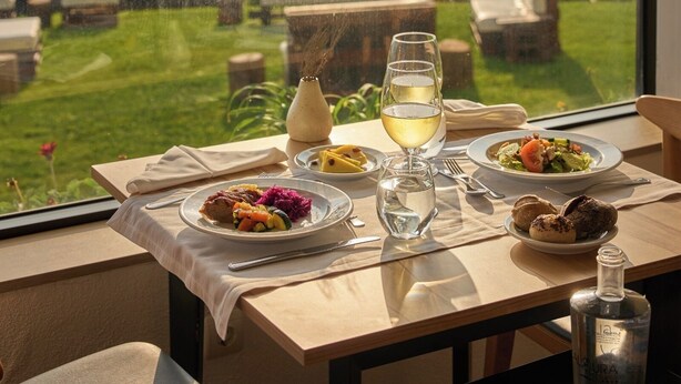 A dining table with plates of food, a bottle of water, and a glass of wine, set against a scenic view of the sea and cliffs. - Caloura Hotel Resort (Lagoa)