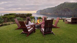 Two people sitting on wooden chairs by a fire pit with a view of a lake and a hilly landscape. - Caloura Hotel Resort (Lagoa)