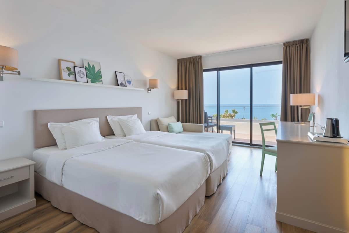 Een hotelkamer met twee bedden, een bureau, een stoel en uitzicht op zee.