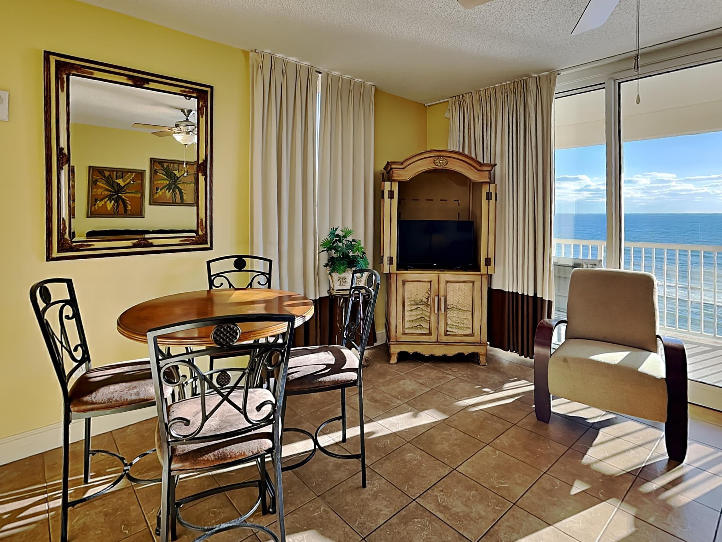 A room with a dining table, chairs, a TV cabinet, and a chair by a large window with a view of the ocean.