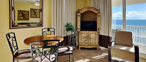 A room with a dining table, chairs, a TV cabinet, and a chair by a large window with a view of the ocean.