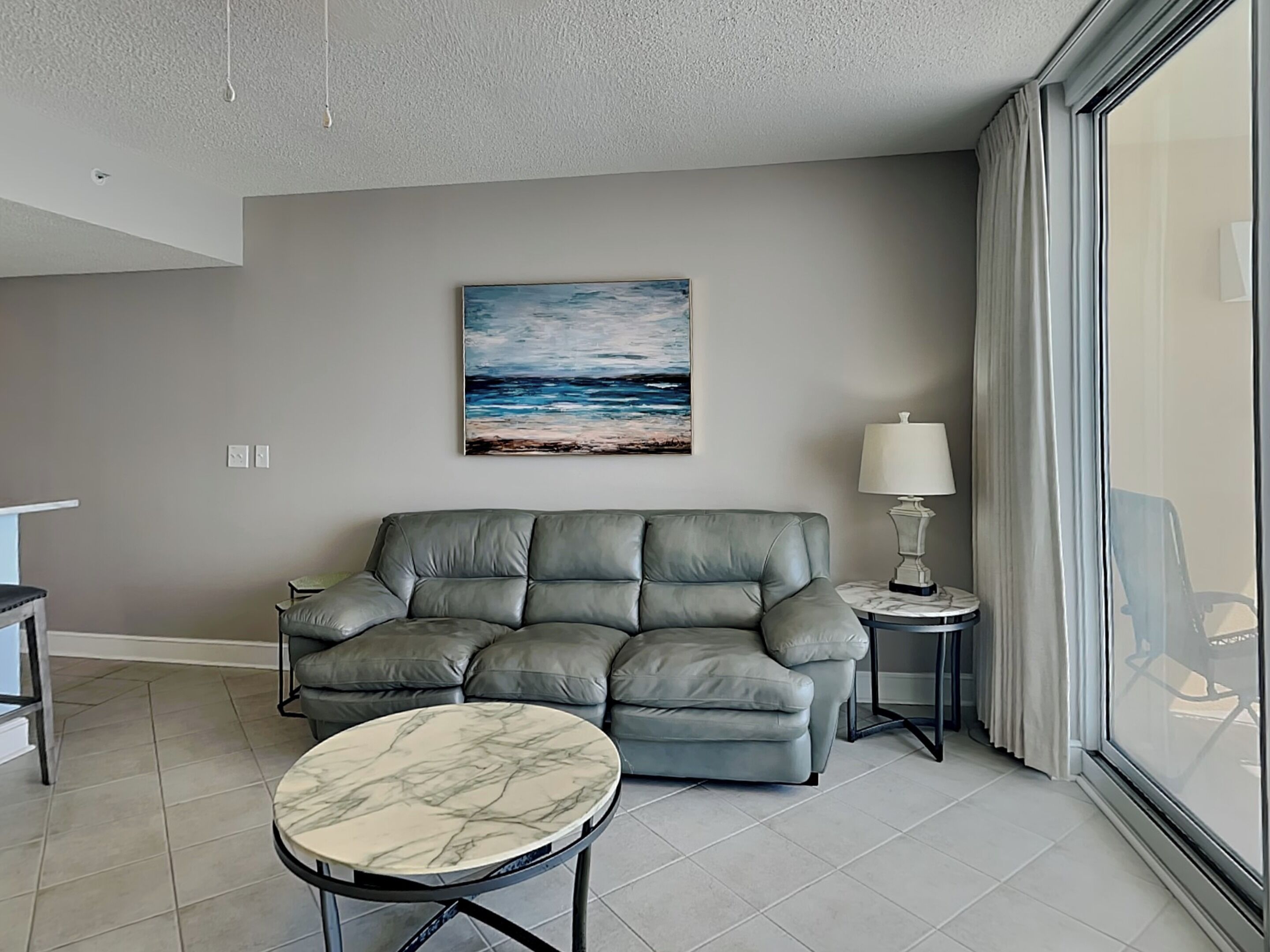 A modern living room with a grey sofa, a marble coffee table, a painting of a beach scene, a lamp, and a view of the outdoors through a large window.