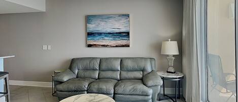 A modern living room with a grey sofa, a marble coffee table, a painting of a beach scene, a lamp, and a view of the outdoors through a large window.
