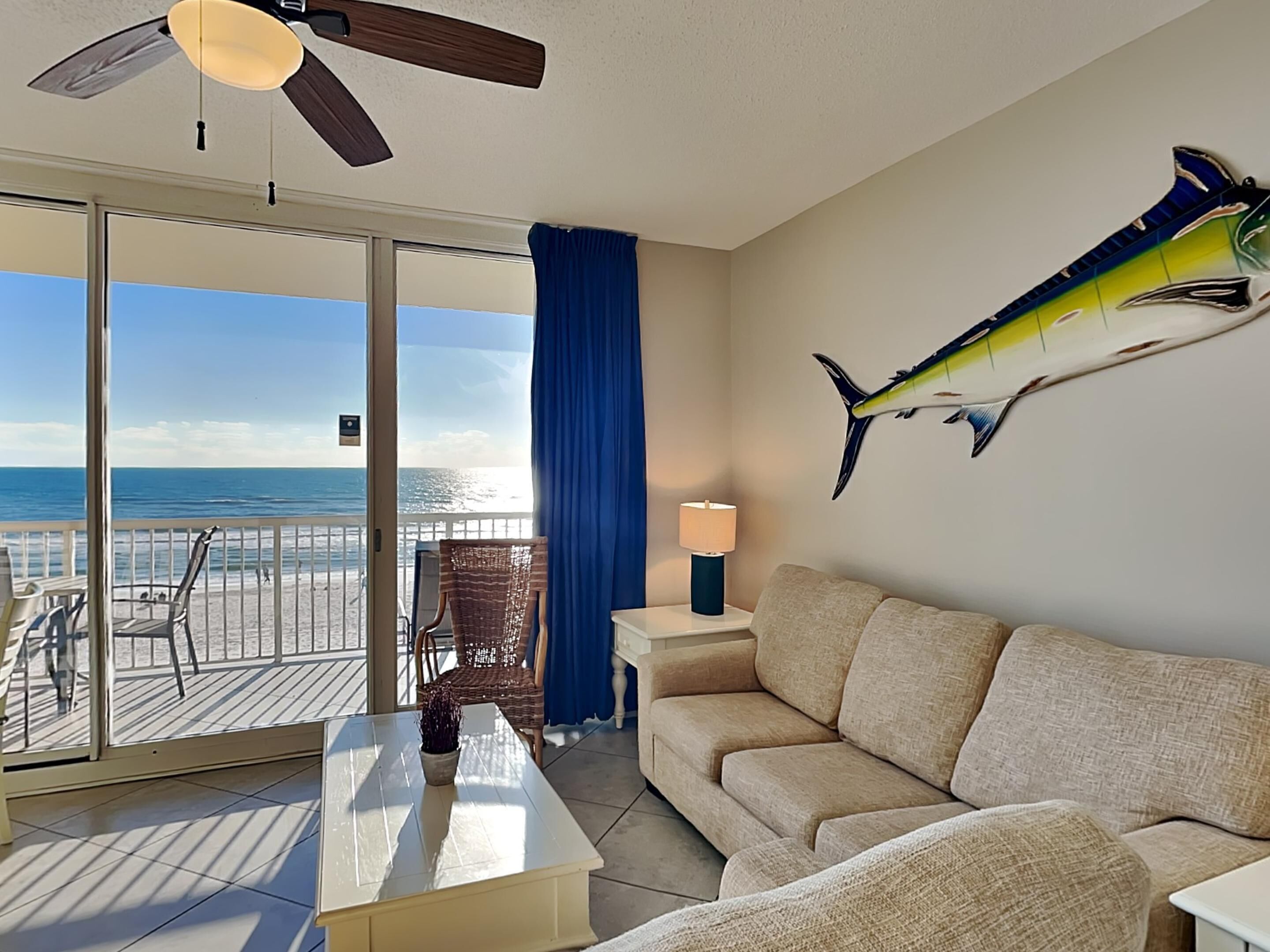 A room with a balcony overlooking the beach, a ceiling fan, a sofa, a table, and a lamp.