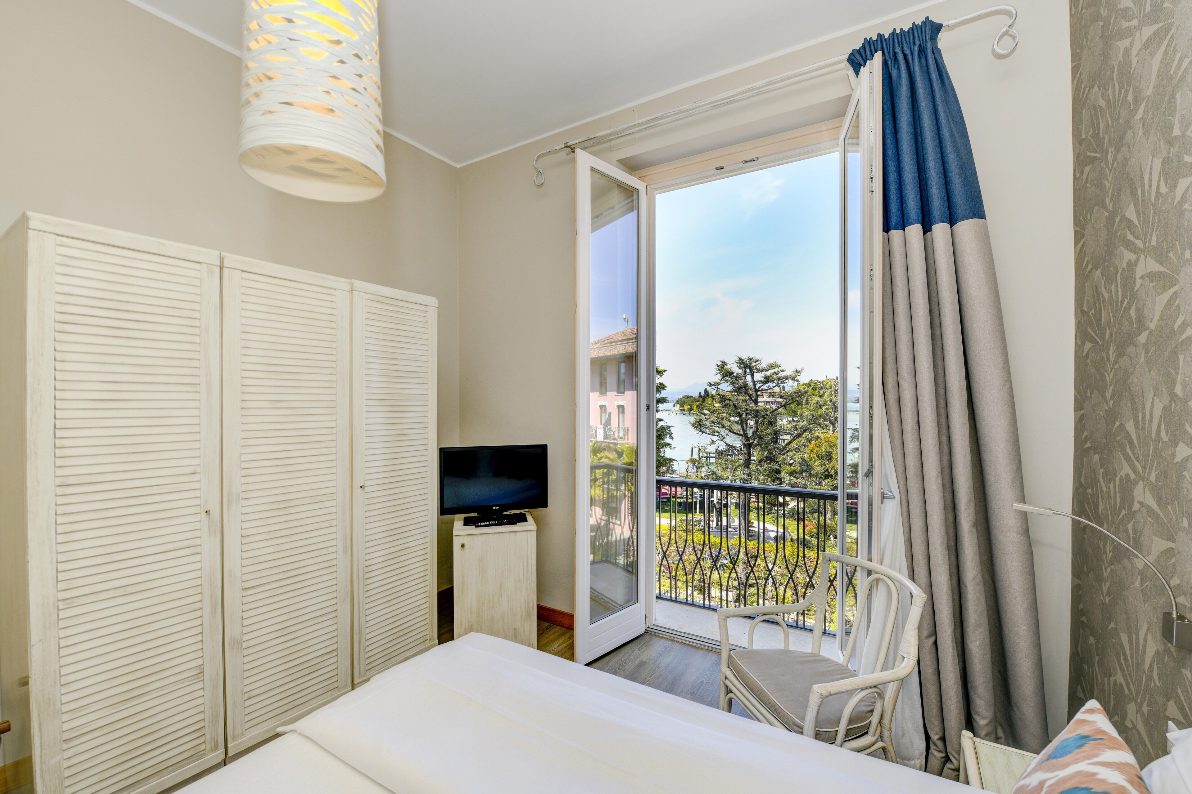 A bedroom with a bed, a TV on a stand, a balcony with a chair, and a view of the outdoors.