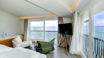 Una habitación de hotel con cama, televisión, balcón con vista al mar y baño con bañera y ducha.