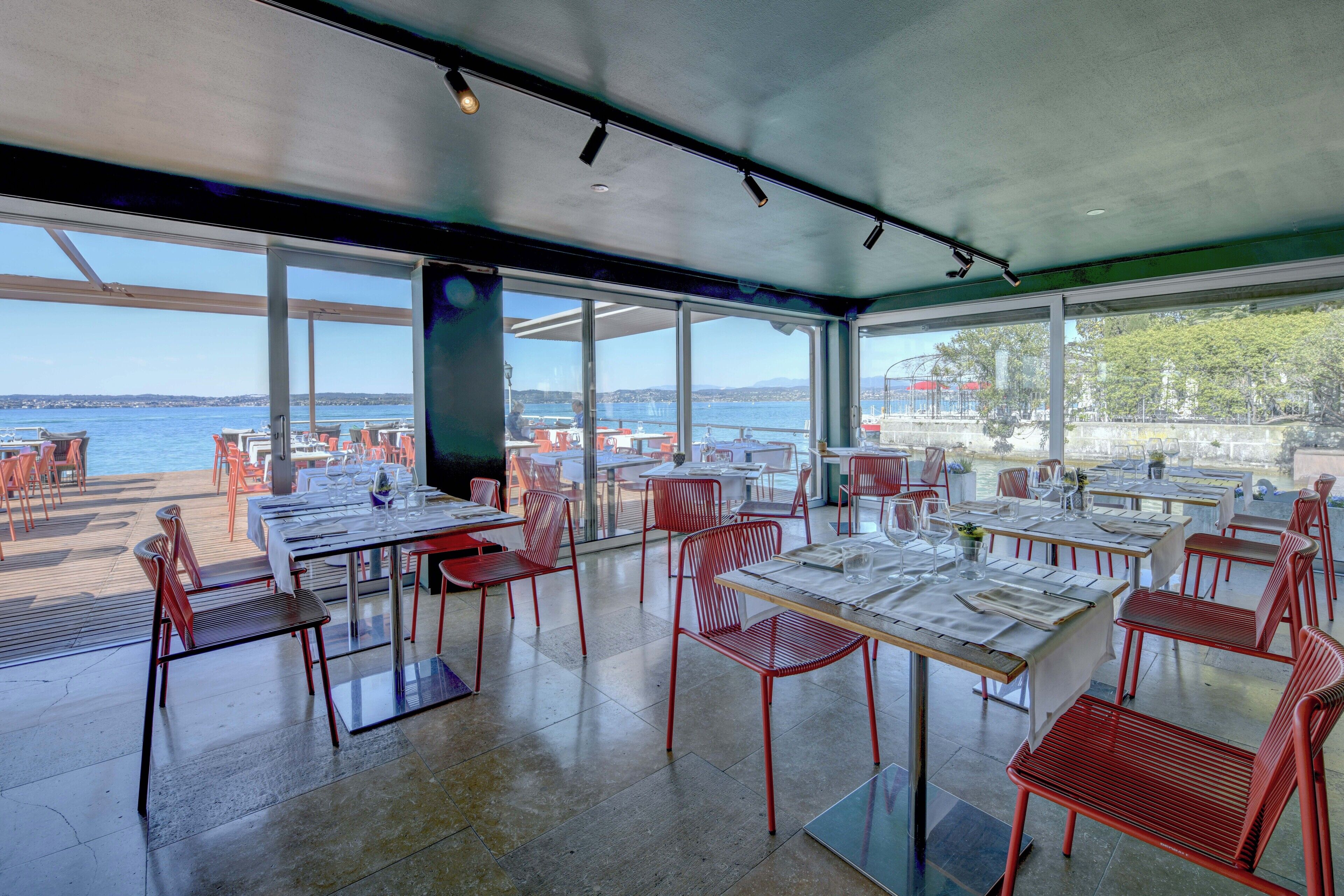 Un espace extérieur aménagé pour les repas, avec des tables et des chaises, de grandes fenêtres et une vue sur l’eau.