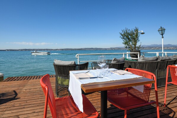 A wooden deck with a table set for two, outdoor seating, a potted plant, and a view of the sea with a boat in the distance.