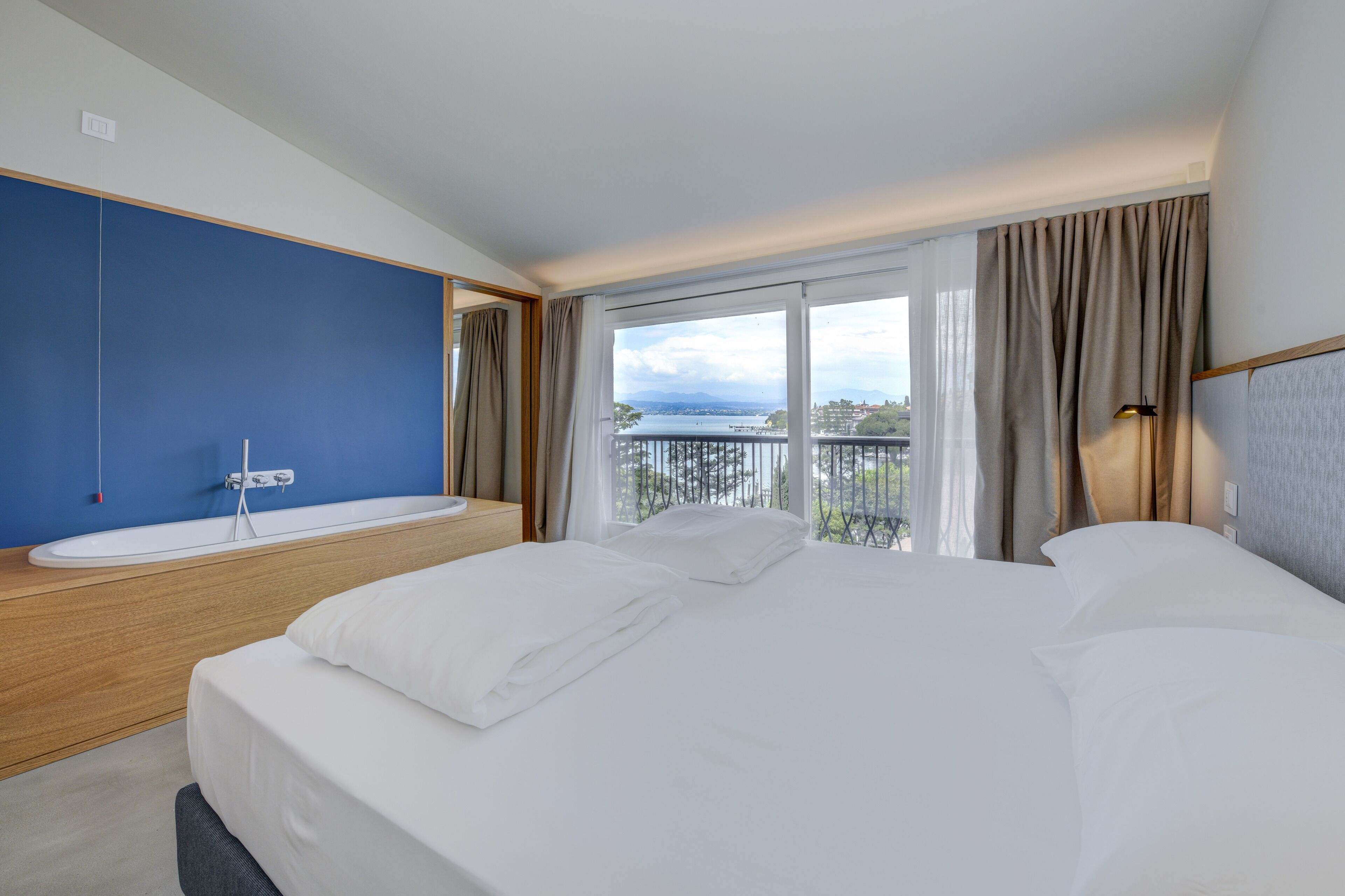 Ein modernes Hotelzimmer mit einem großen Bett, einer integrierten Badewanne und einem Balkon mit Stadtblick.