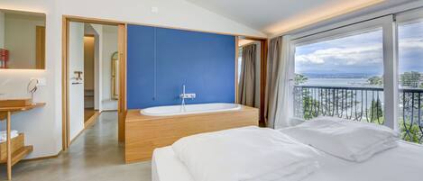 A modern hotel room with a large bed, a bathtub, and a balcony with a view of the sea.