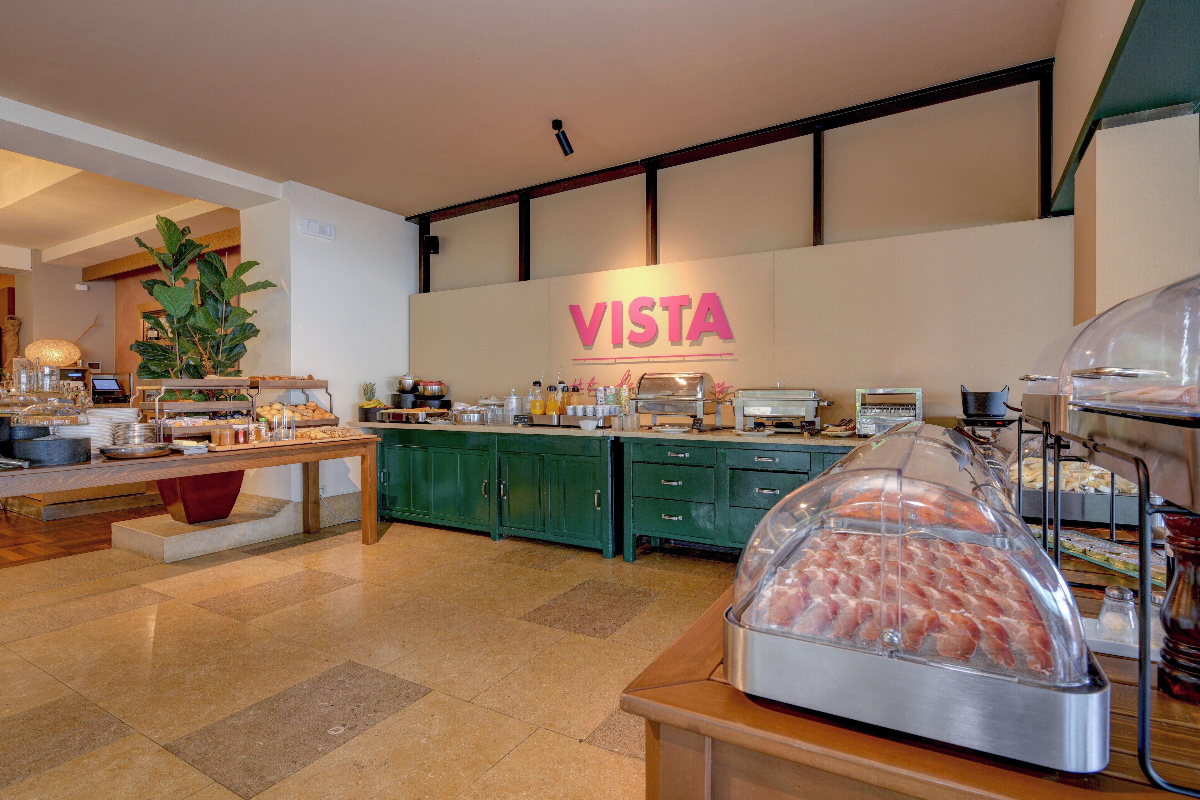 Een ontbijtbuffet met een 'VISTA' bord, groene kastjes en diverse soorten eten.