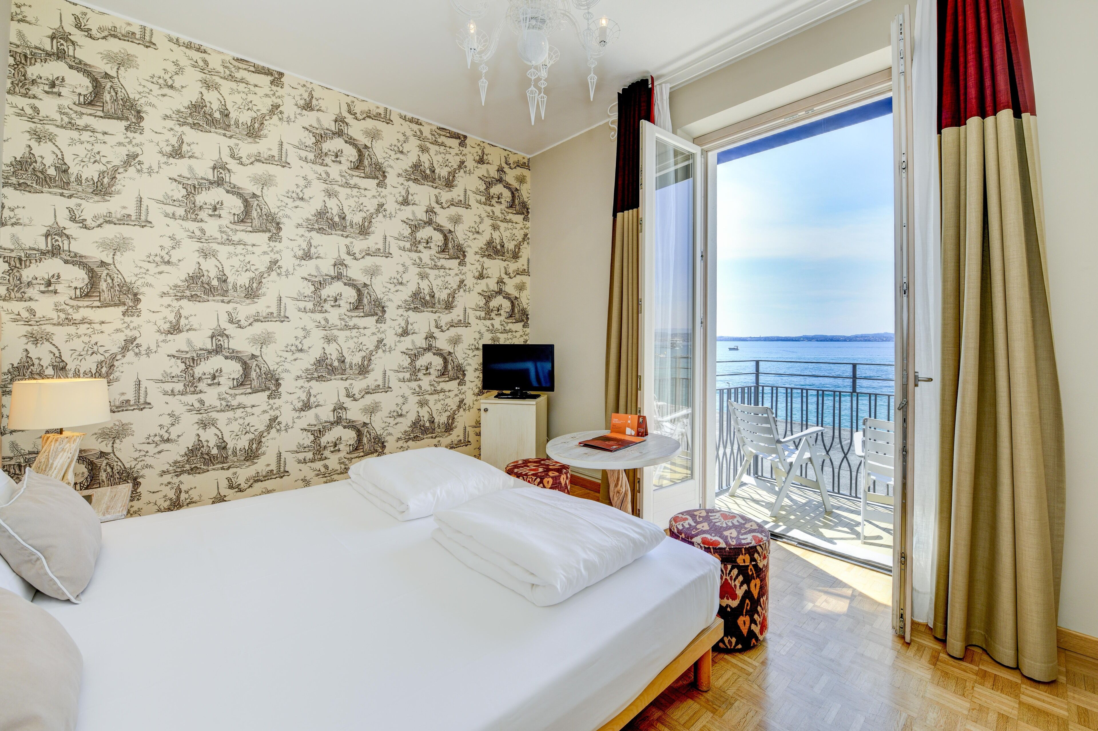 A hotel room with a bed, a television, a small table, a chair, and a balcony with a view of the sea.