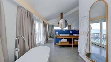 Ein modernes Hotelzimmer mit einer großen Badewanne, einem Schminktisch mit Spiegel, einem Holfnachtisch und Meerblick durch ein großes Fenster.