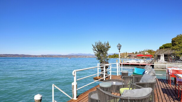 A wooden deck with wicker furniture overlooking a body of water, a lamp post, and a distant view of a cityscape. - Hotel Flaminia (Sirmione)