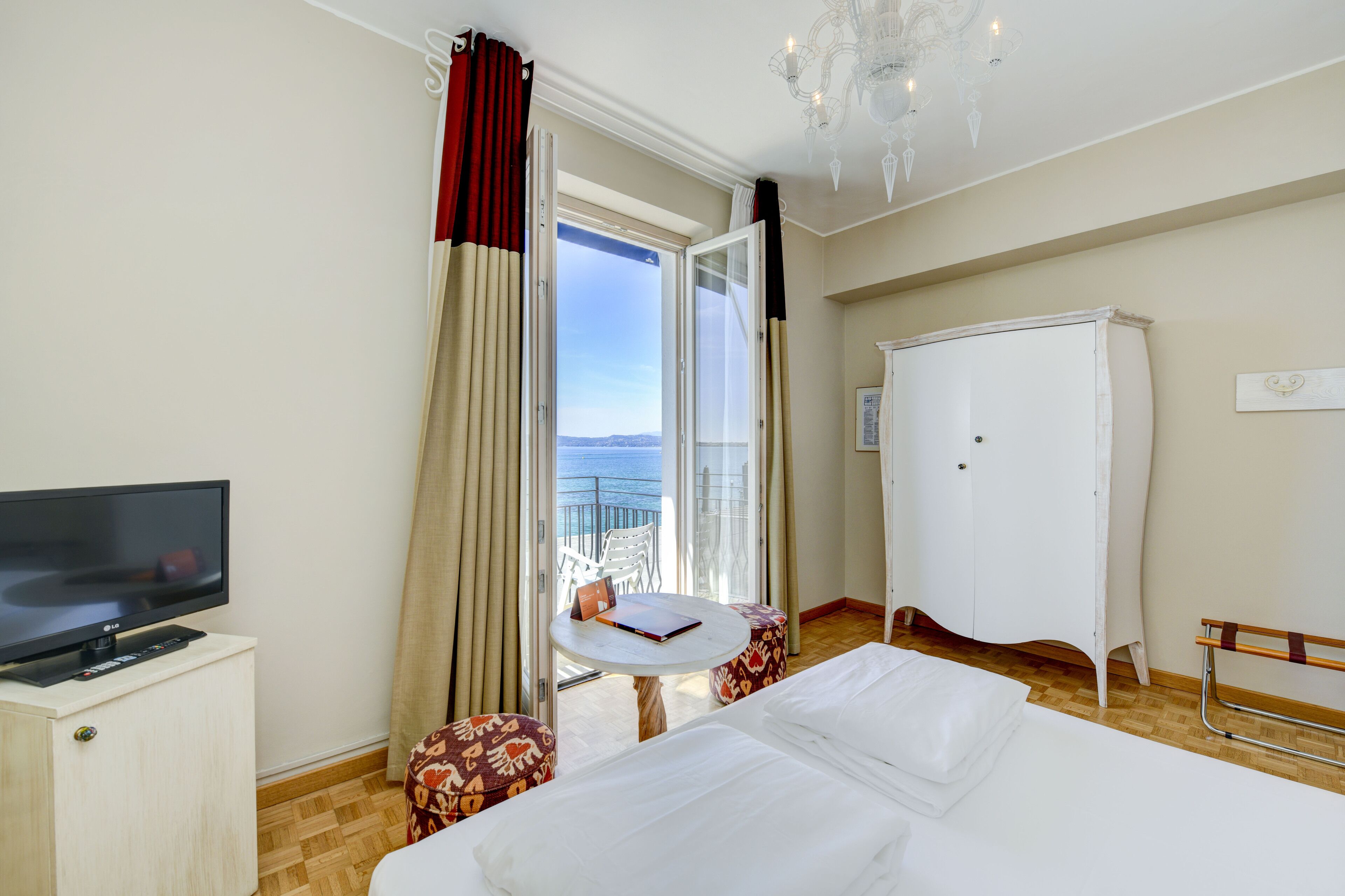 A hotel room with a bed, a TV, a small table, a chair, a wardrobe, and a balcony with curtains.