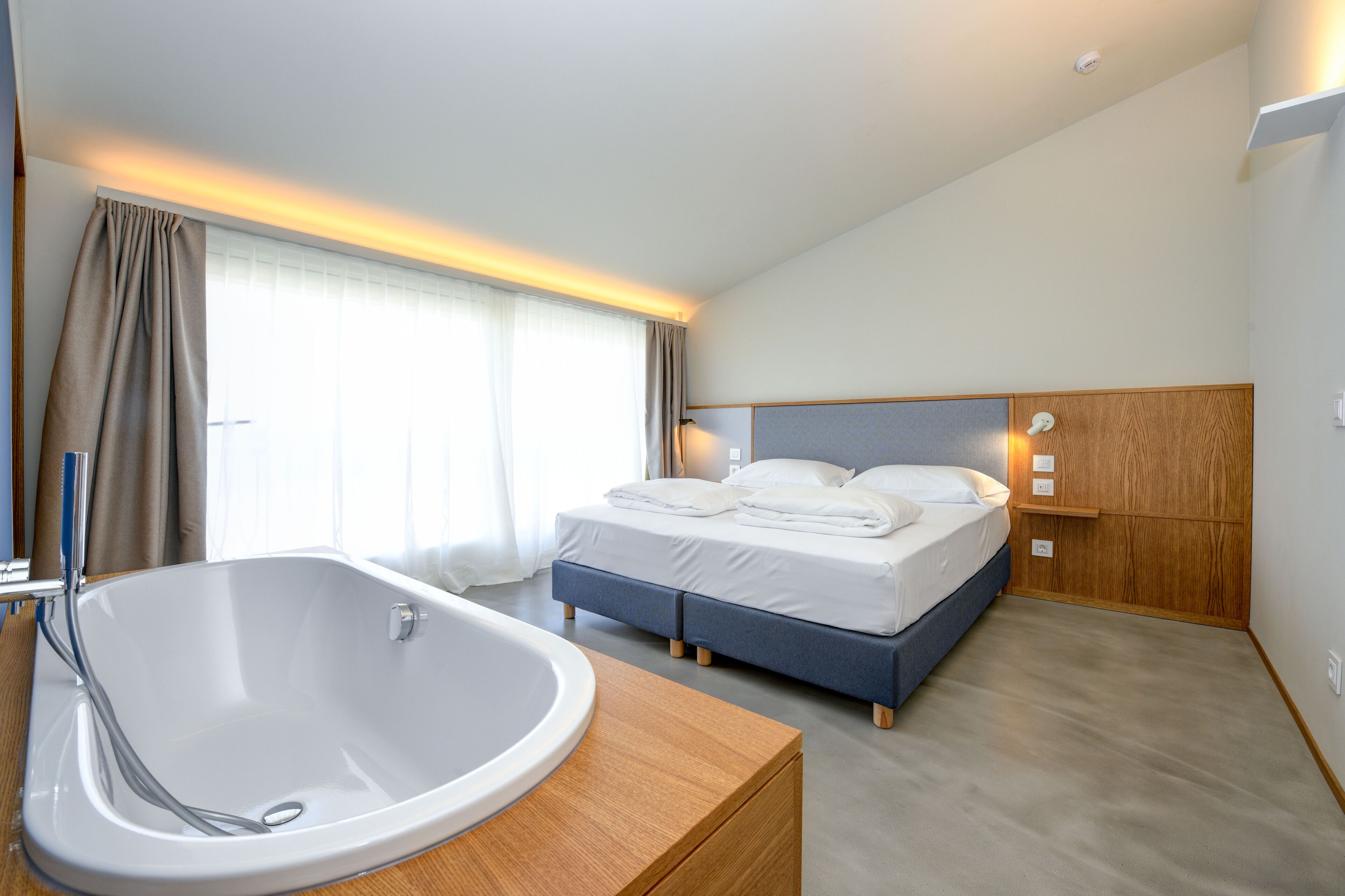 A modern hotel room with a large bathtub, a neatly made bed, and a wooden headboard.