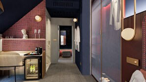 Un pasillo moderno de hotel con pared de diseño rojo, una zona de estar con vitrina de cristal, y pared azul con espejo y perchero.