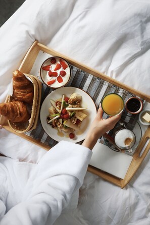 Ein Frühstücksbrett mit Croissants, Joghurt mit Erdbeeren, einer Obst- und Nussplatte, einem Glas Orangensaft und einer Kaffeetasse auf einem Bett mit weißer Bettwäsche.