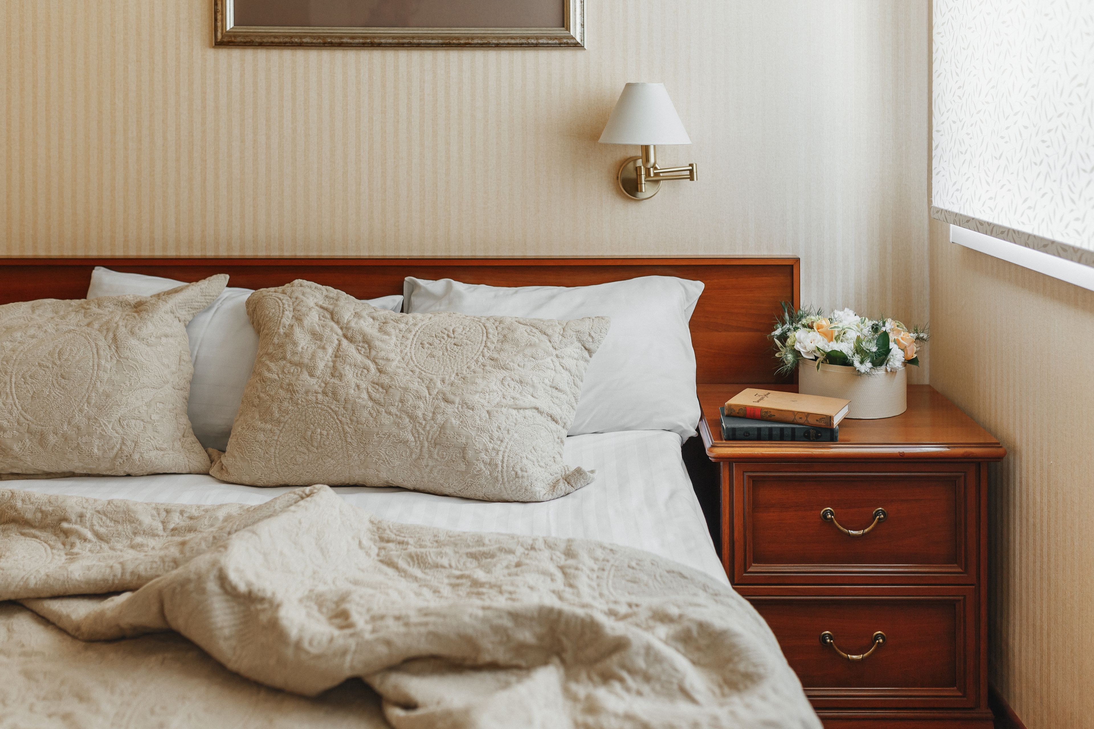 A neatly made bed with a white comforter and decorative pillows, a bedside table with a lamp and a vase of flowers, and a wooden nightstand with drawers.