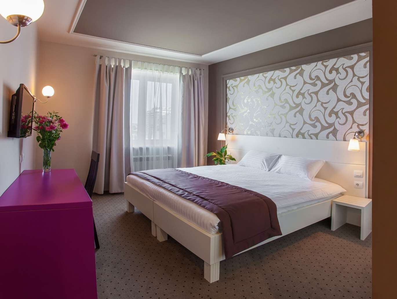 A hotel room with a bed, bedside tables, a desk, a chair, a vase with flowers, and a window with curtains.