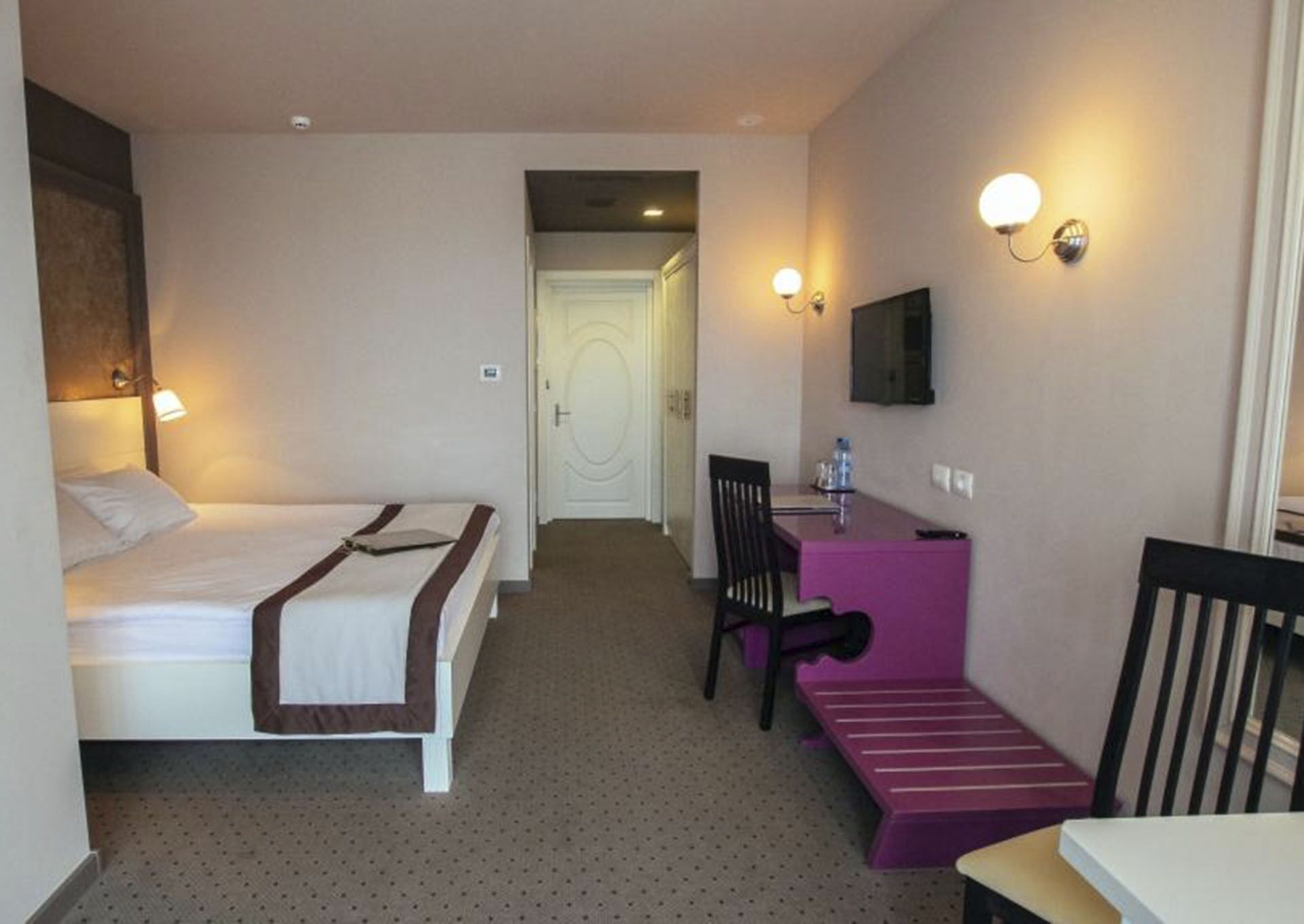 Quarto de hotel com cama, escrivaninha, cadeira, mesinha e televisão na parede.