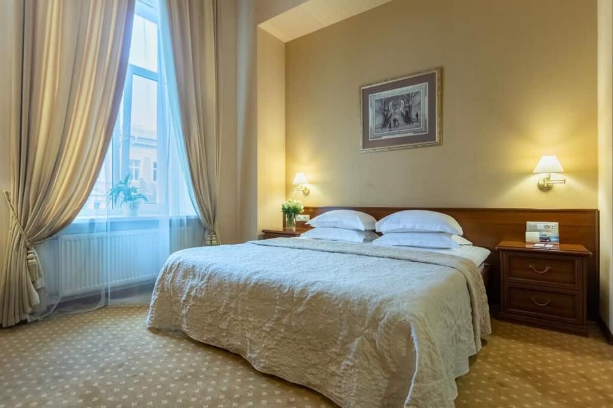 Habitación de hotel con una cama grande, dos mesitas de noche con lámparas, una ventana con cortinas y un cuadro en la pared.
