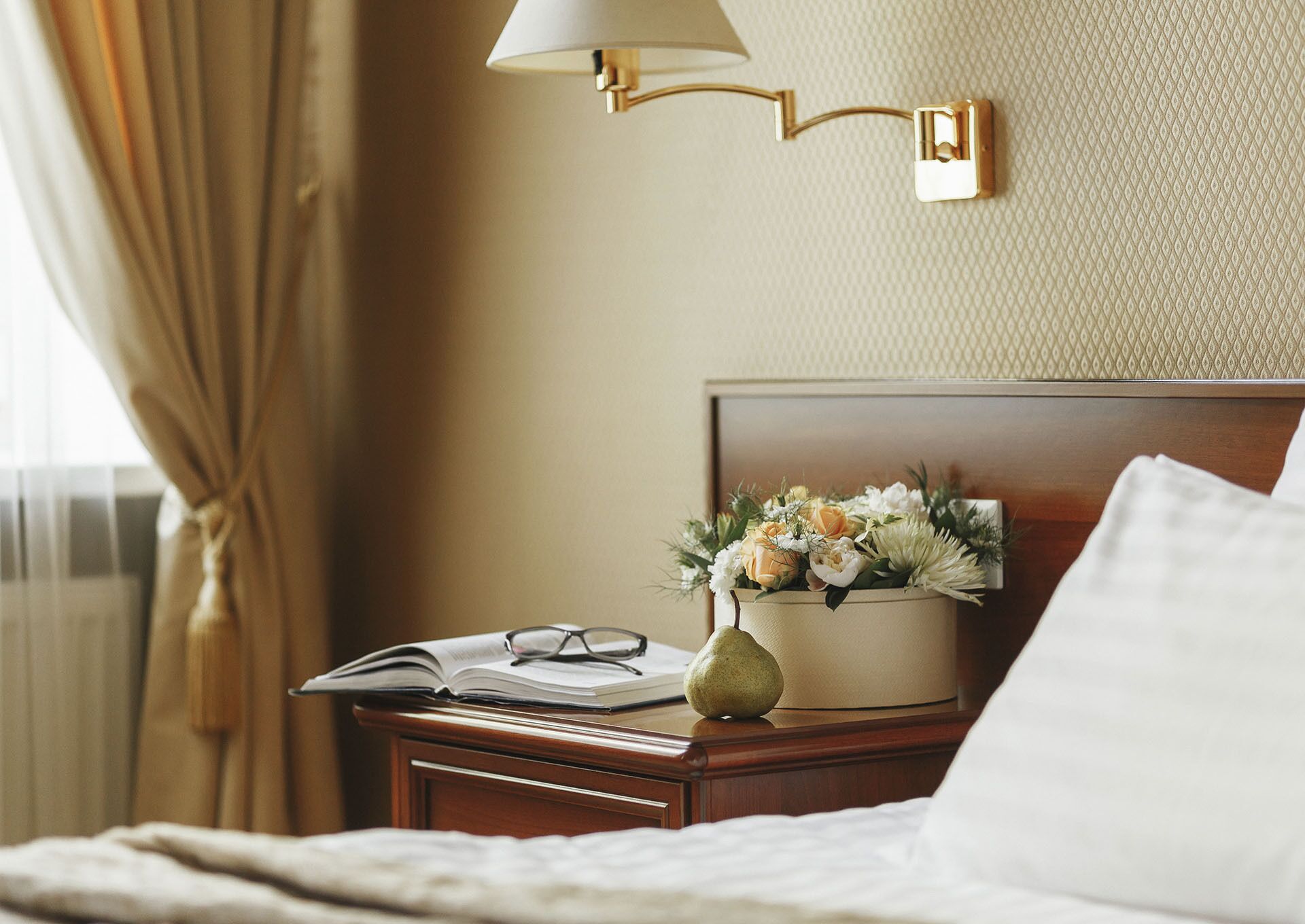 A neatly made bed with a white pillow, a bedside table with a lamp, an open book, and a vase with flowers.