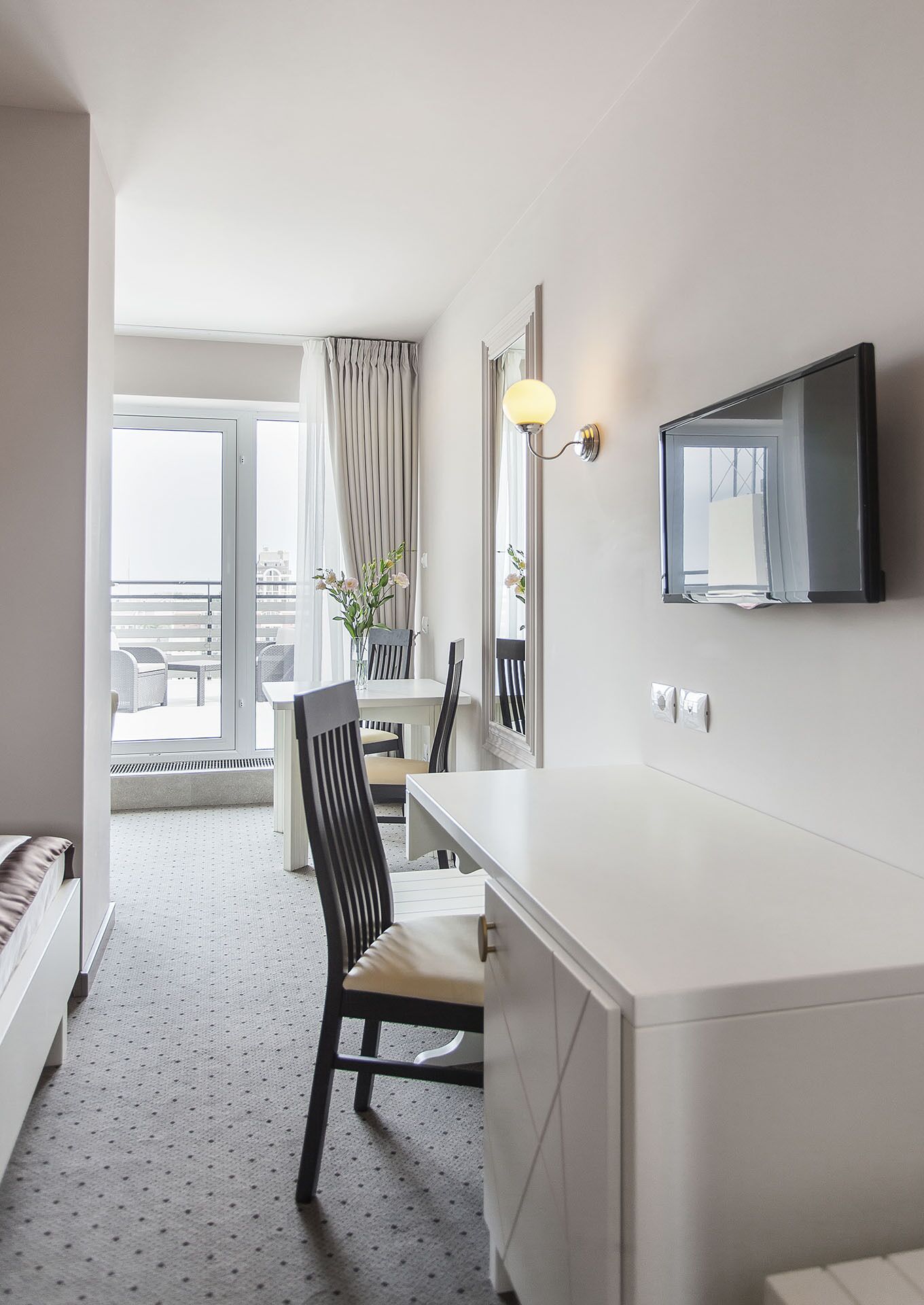 A hotel room with a large window, a dining area with a table and chairs, a white desk with a chair, a flat-screen TV mounted on the wall, and a mirror.