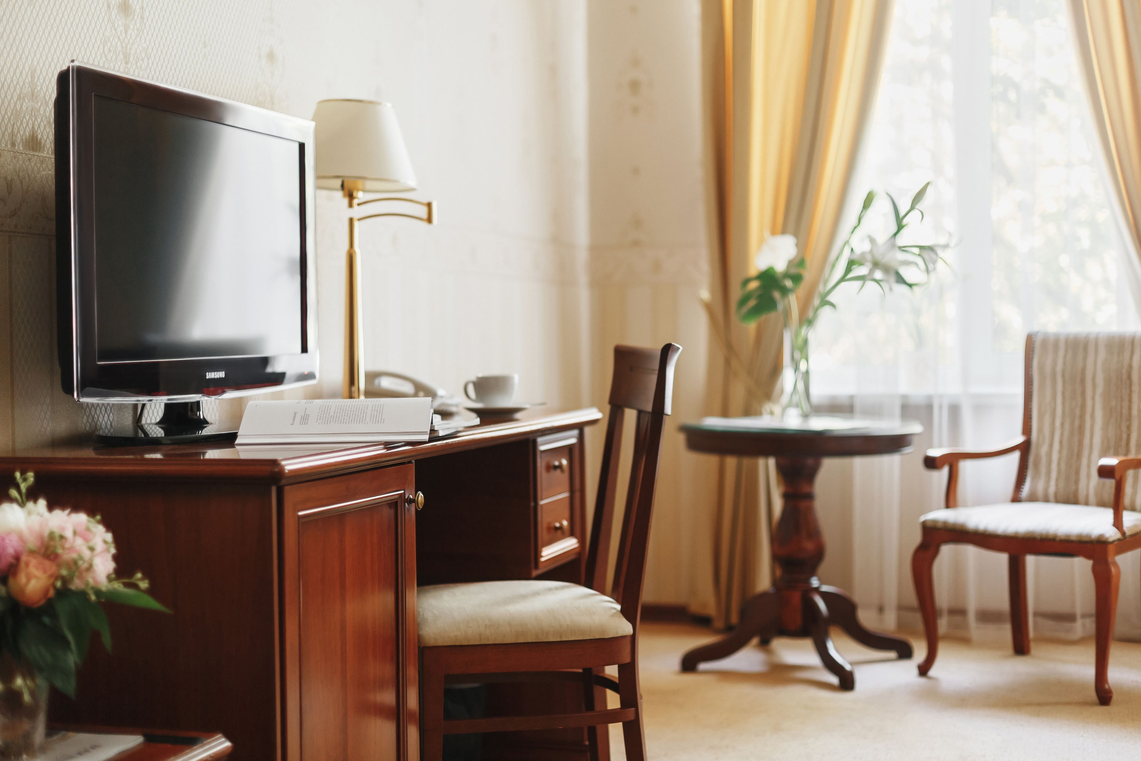 Ein Hotelzimmer mit Schreibtisch aus Holz, einem Flachbildfernseher, einer Lampe, einer Vase mit Blumen und Blick ins Freie durch ein Fenster.