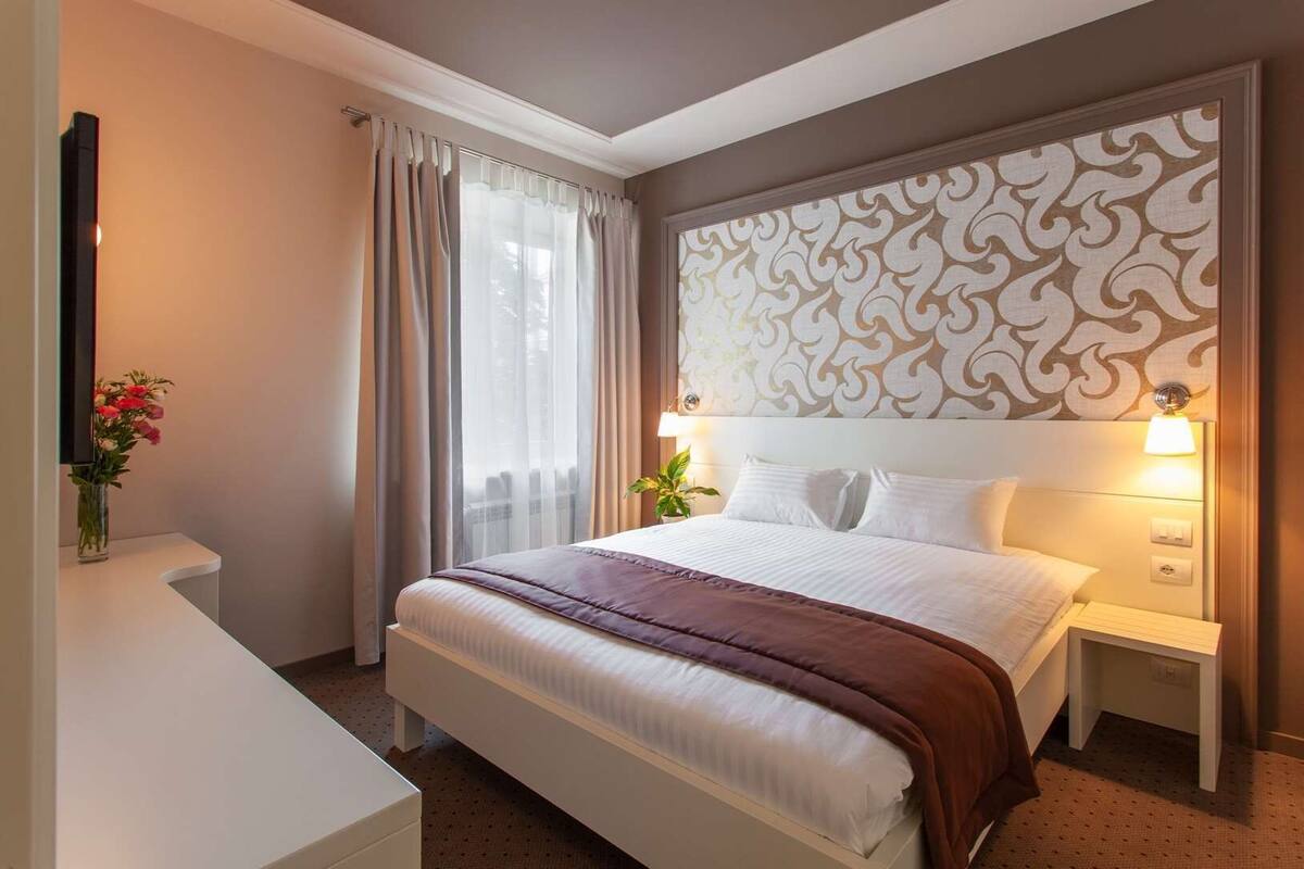 Habitación de hotel con cama, mesitas de noche, lámpara, televisor montado en la pared, ventana con cortinas y cabecera decorativa.