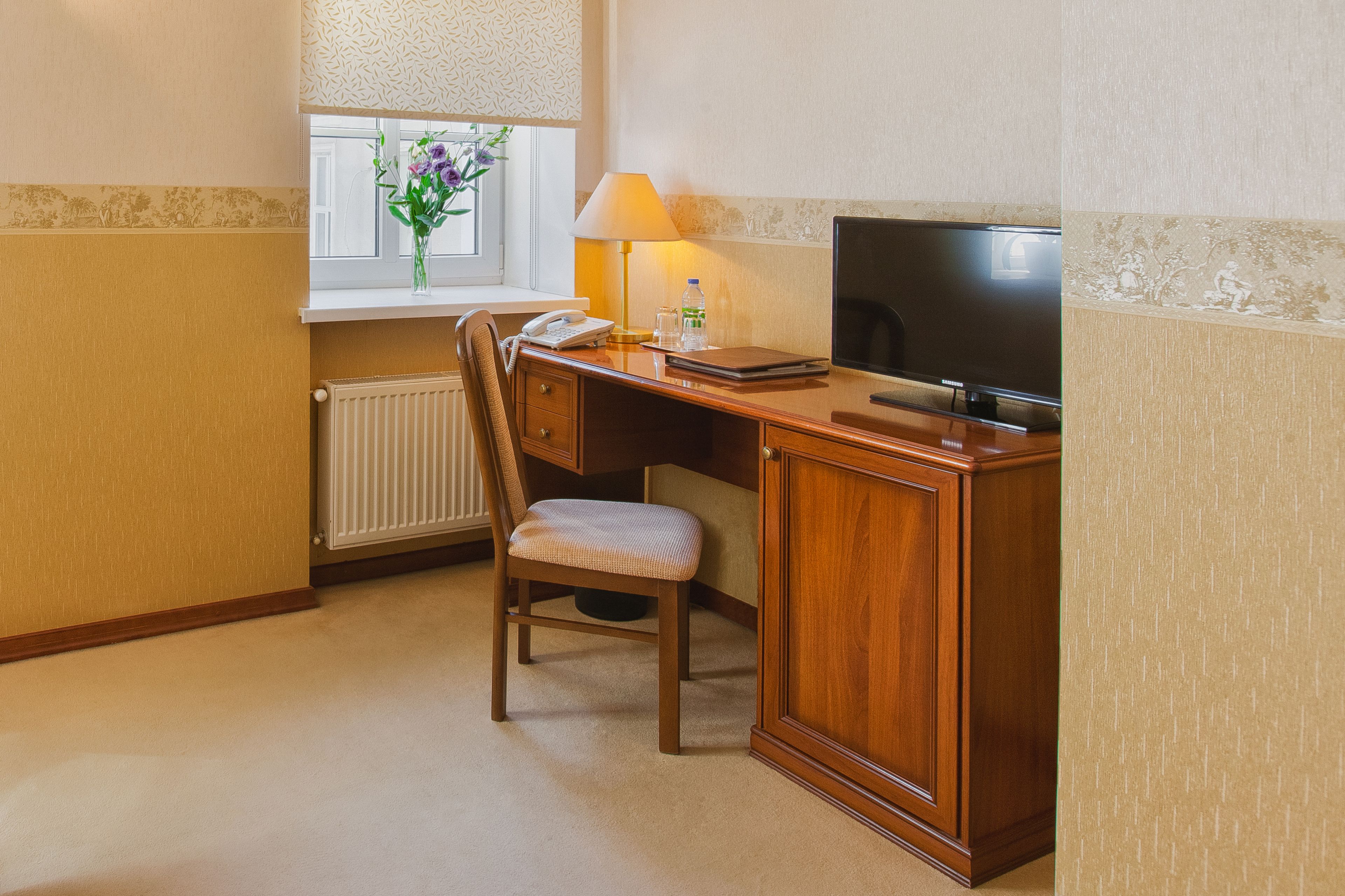 A hotel room with a wooden desk, a chair, a television, a lamp, a telephone, and a window with a vase of flowers.