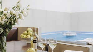 Uma mesa com uma garrafa de vinho, dois copos, uma maçã e um vaso de flores. Ao fundo, vista de uma piscina.