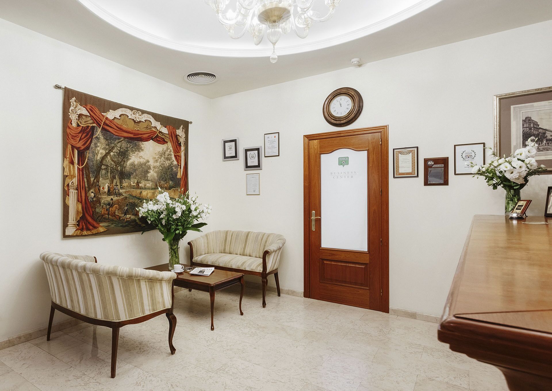 Una habitación con un cuadro clásico grande, una puerta de madera con la etiqueta 'BUSINESS CENTER' y varios certificados enmarcados en la pared.