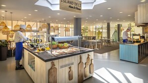 Un restaurant moderne avec une enseigne indiquant « Island Deli : jambon et fromage », comprenant un comptoir central où sont exposés les plats et un chef qui prépare les aliments.