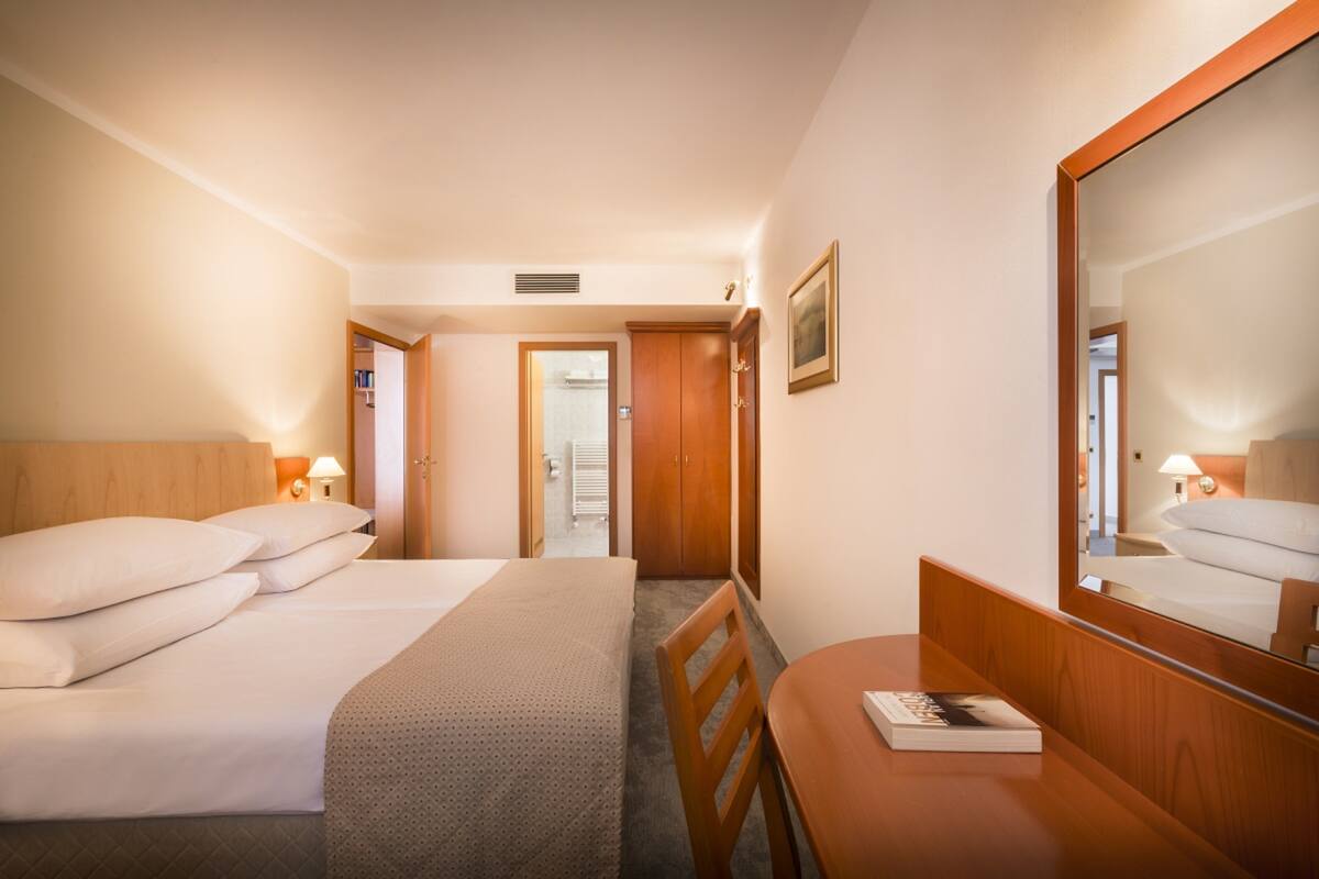 A hotel room with a bed, a desk, and a mirror. There is a door leading to another room.