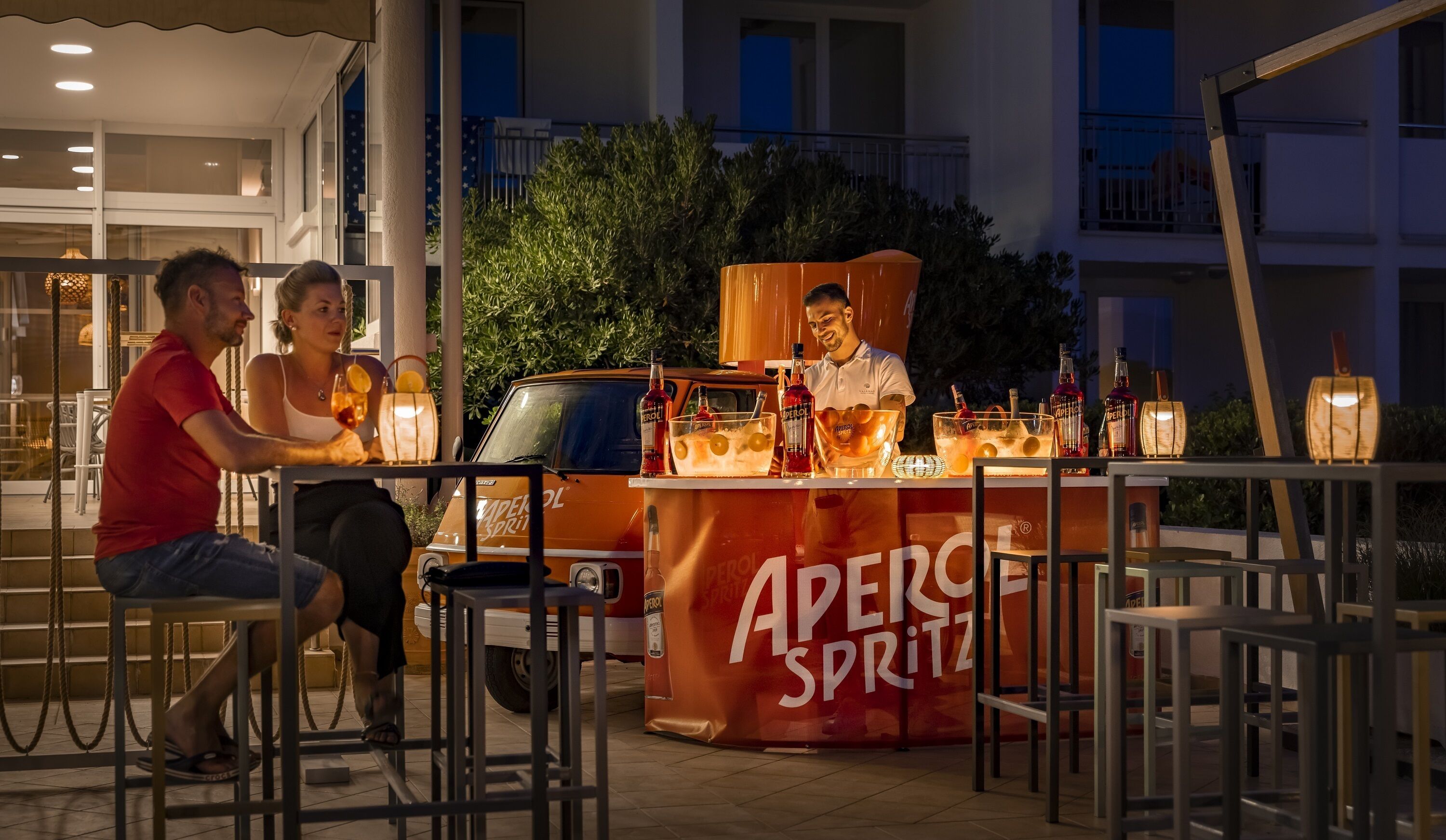 An outdoor bar area with a branded beverage stand, seating, and a vehicle in the background.