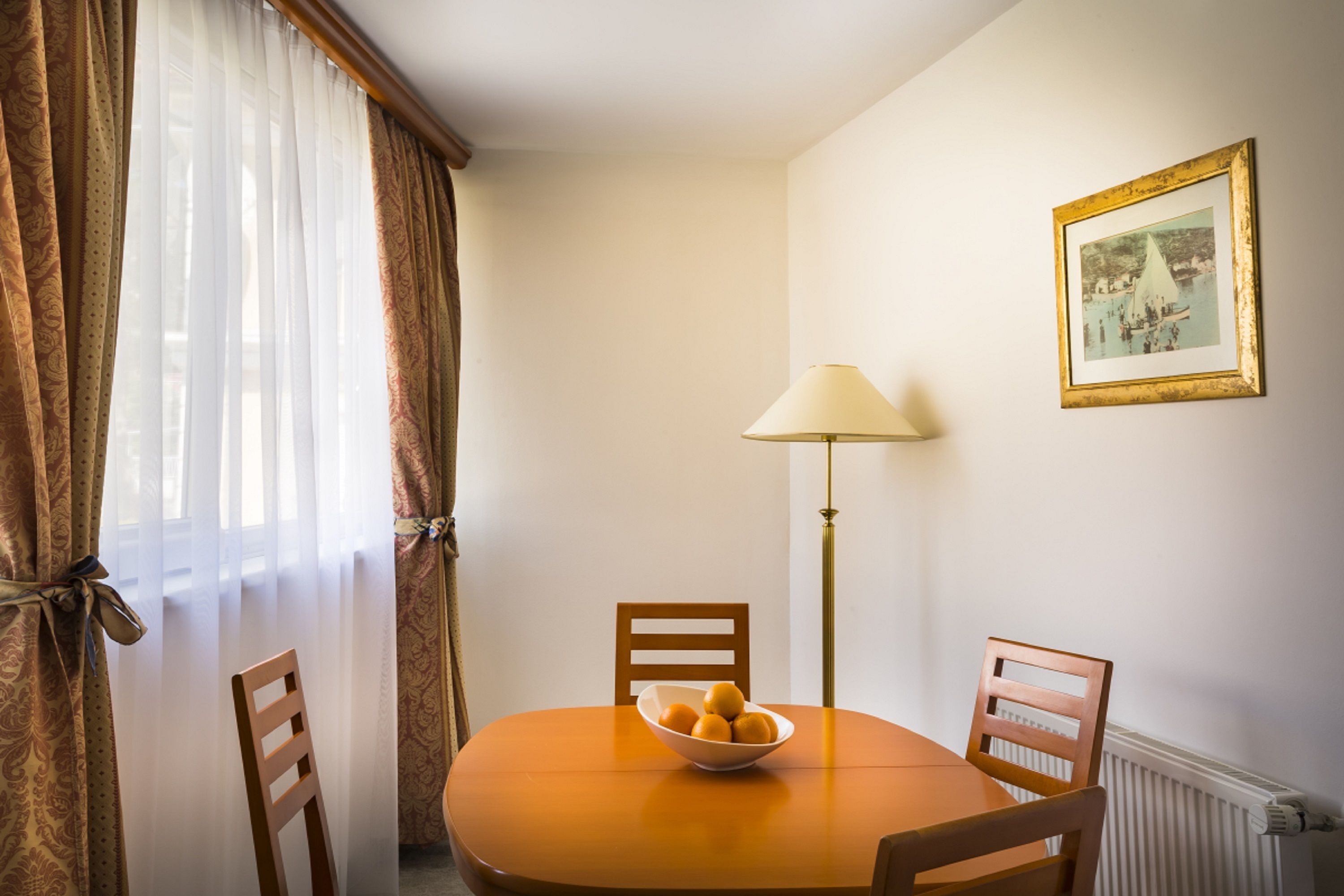 A dining area with a round table, two chairs, a bowl of fruit, a lamp, and a framed picture on the wall.