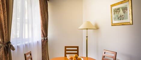A dining area with a round table, two chairs, a bowl of fruit, a lamp, and a framed picture on the wall.
