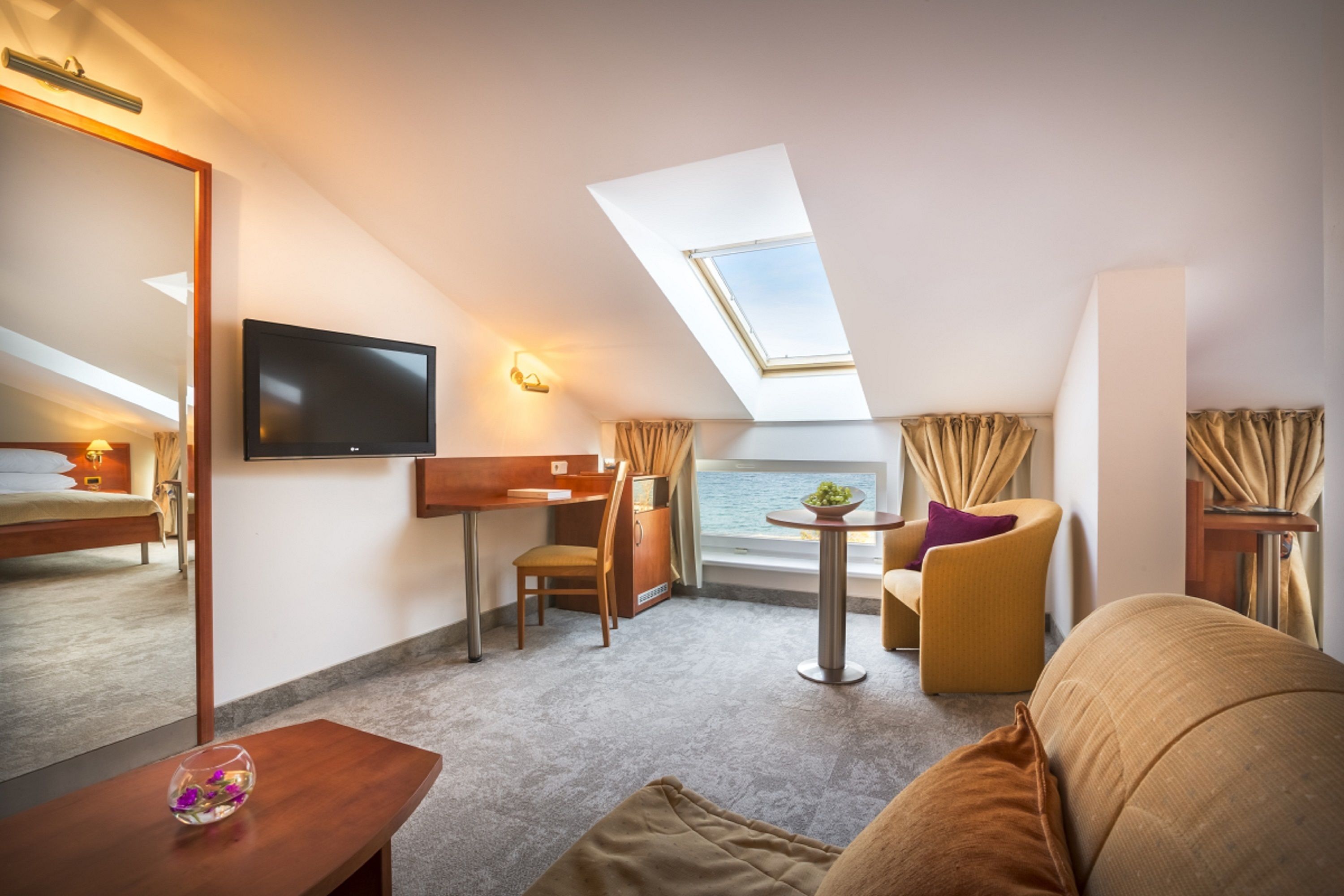 Ein modernes Hotelzimmer mit einem großen Bett, einem Schreibtisch, einer Couch, einem kleinen Tisch mit einer Vase, einem Fernseher und Meerblick durch ein Oberlicht.