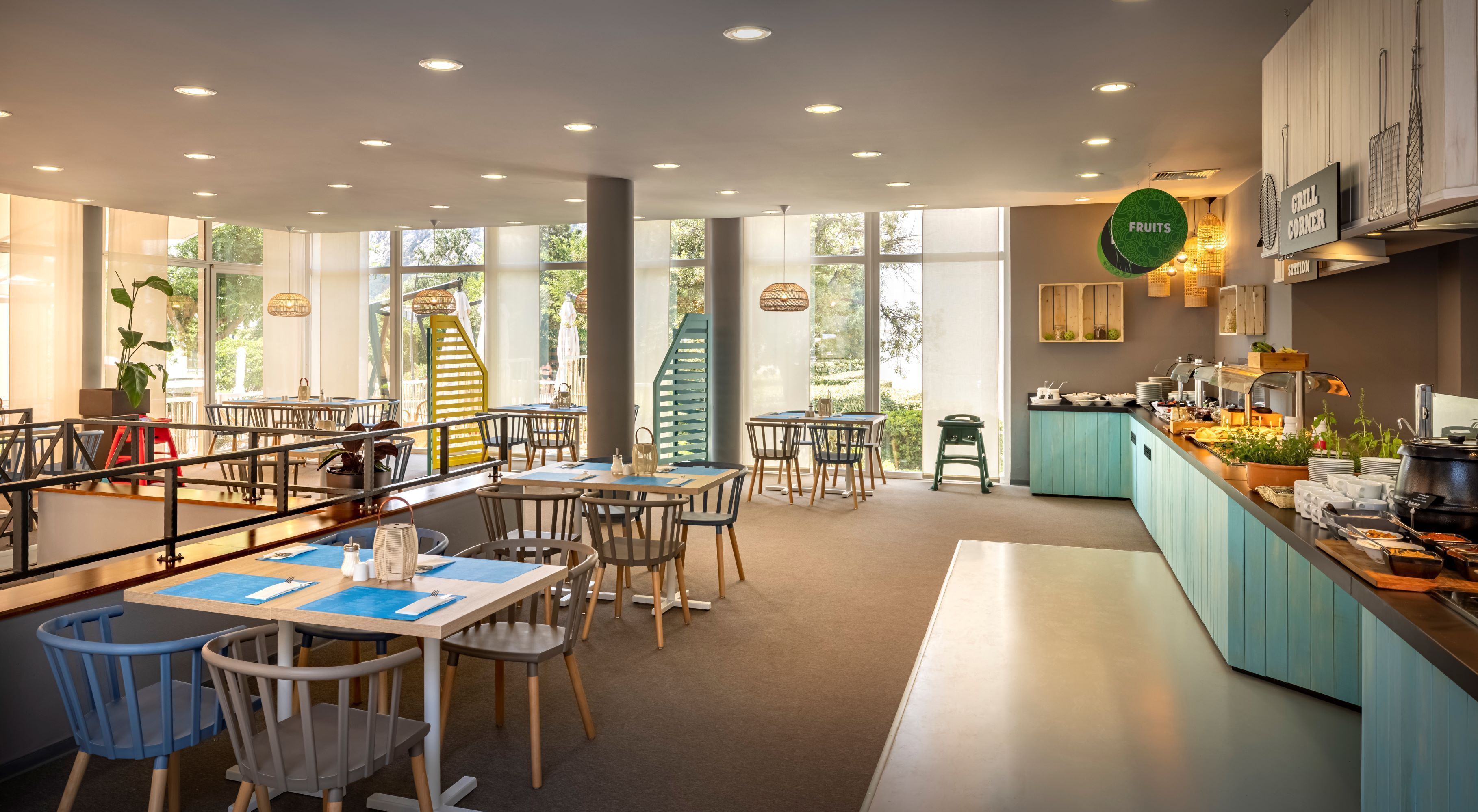 Ein modernes Restaurant mit einer Buffetinsel, Sitzgelegenheiten mit Tischen und Stühlen sowie großen Fenstern mit Blick ins Freie.