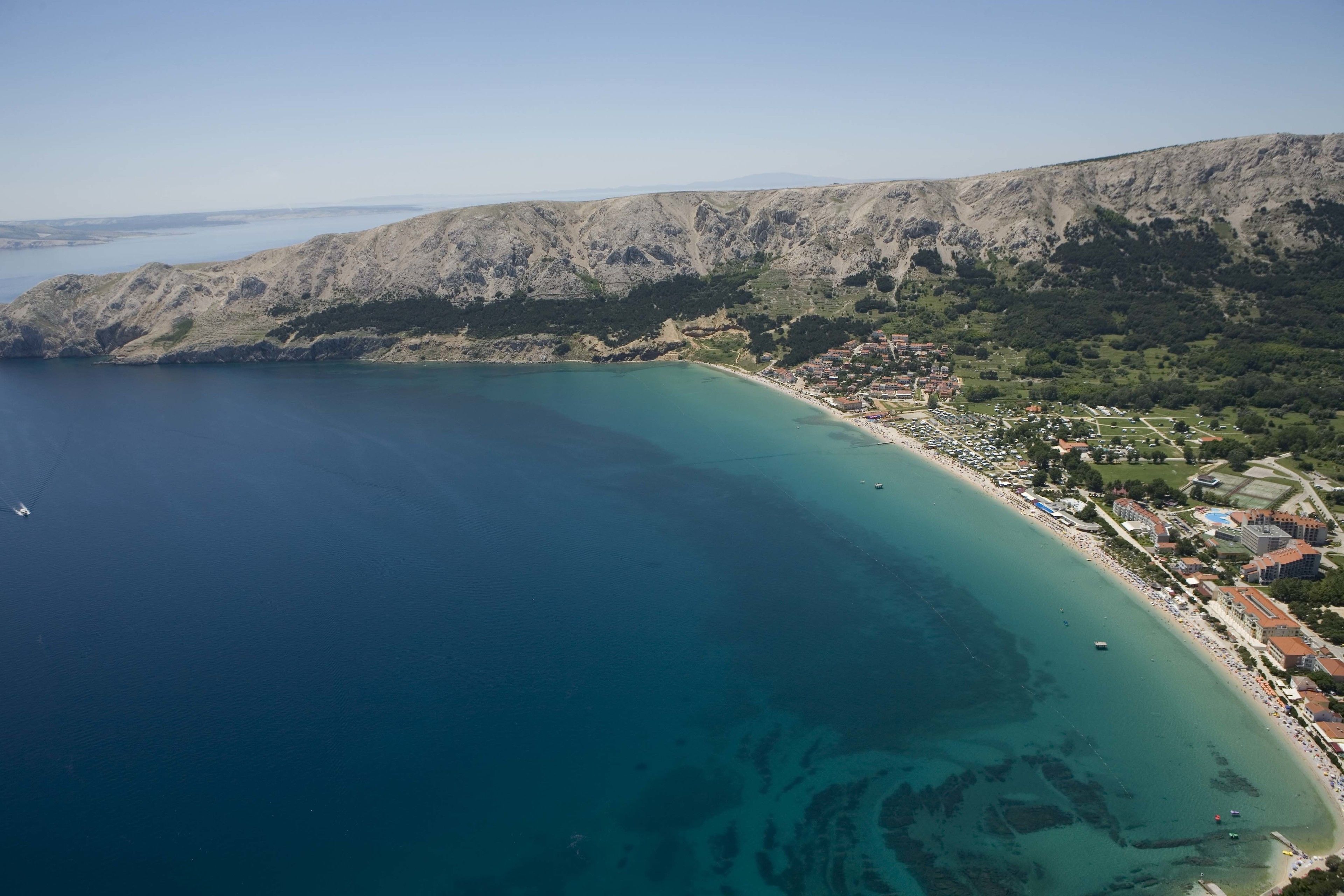 Vista aérea de un pueblo costero con mar azul cristalino, montañas al fondo y una combinación de edificios residenciales y comerciales.