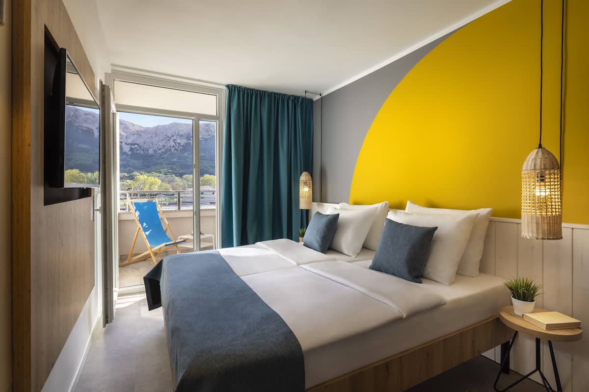 A modern hotel room with a large bed, a view of mountains through the window, and a bright yellow accent wall.