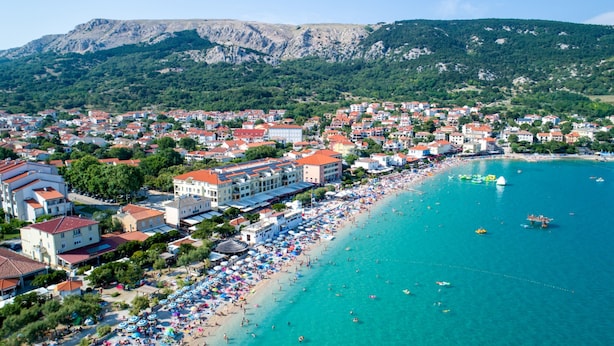 A coastal town with a beach, buildings, and mountains in the background. - Sunny Baška Hotel by Valamar, ex Corinthia (Baska)