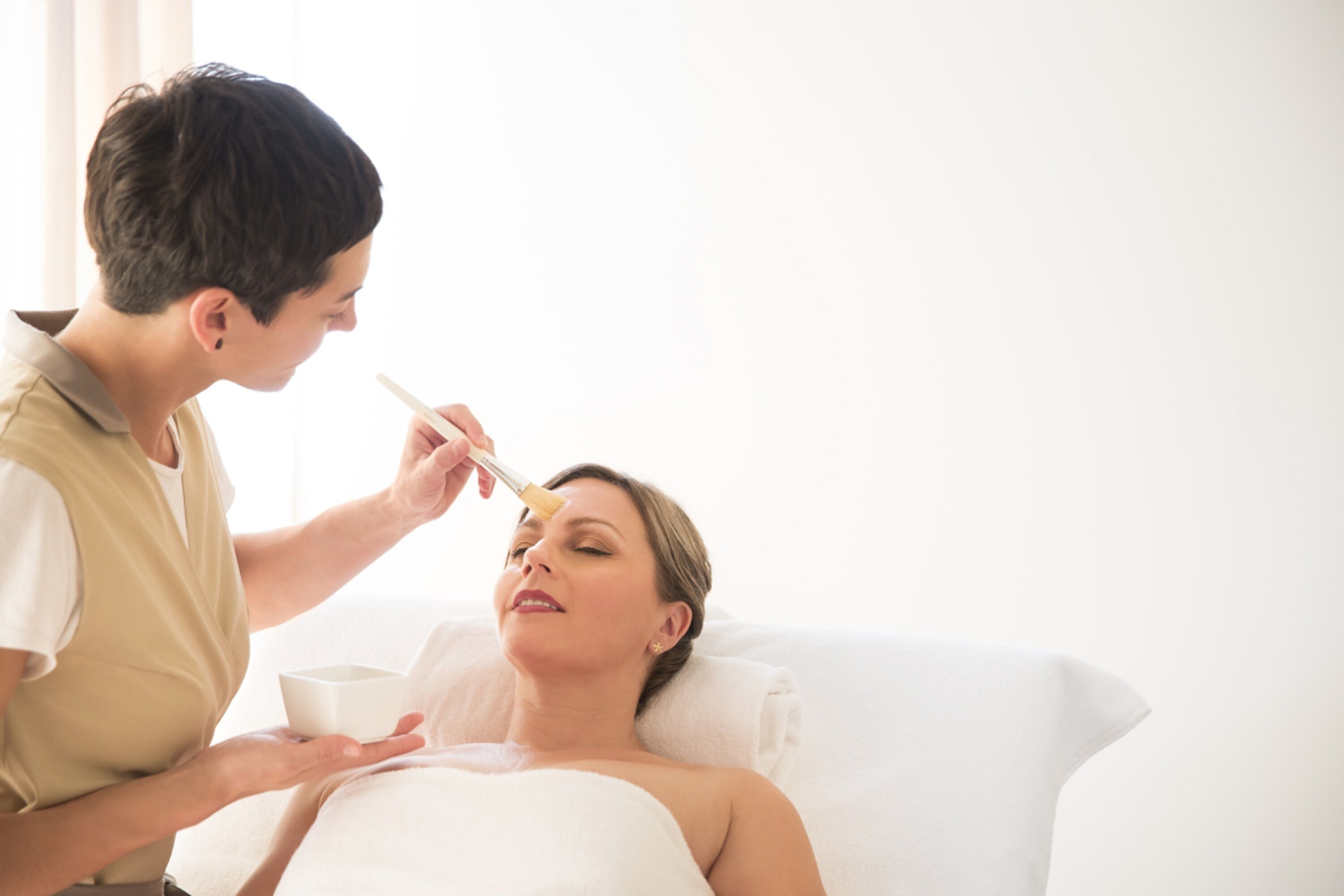 Una persona recibiendo un tratamiento facial en una habitación con una cama blanca y una ventana al fondo.