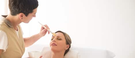 A person receiving a facial treatment in a room with a white bed and a window in the background.