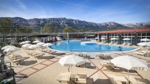 A pool area with lounge chairs and umbrellas, a fountain, and mountains in the background. - Sunny Baška Hotel by Valamar, ex Corinthia (Baska)