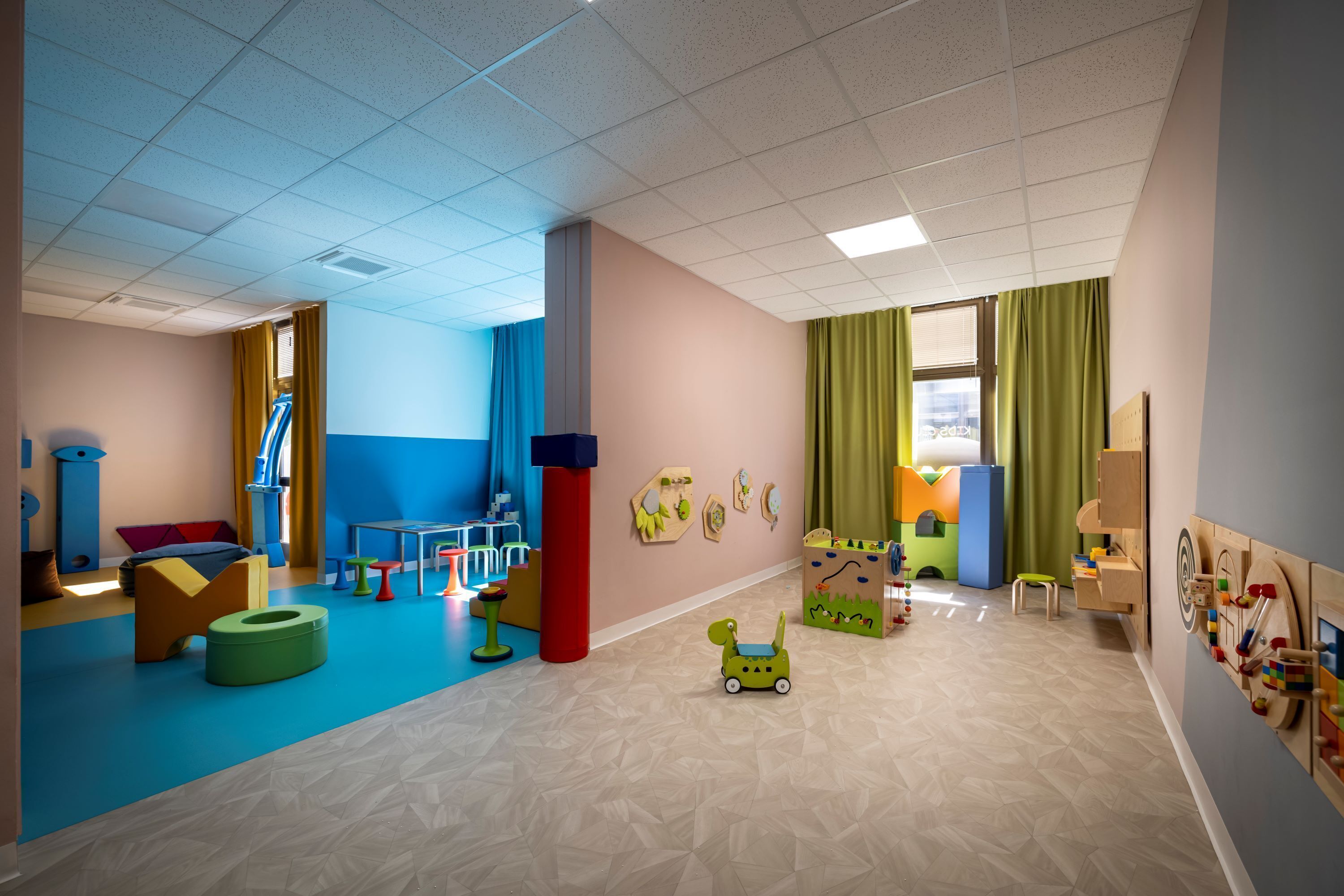Una sala de juegos de colores vivos, con piso azul y beige, un ventanal con cortinas verdes, y diversos juguetes y muebles infantiles.