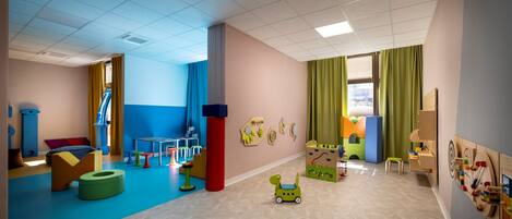Una sala de juegos de colores vivos, con piso azul y beige, un ventanal con cortinas verdes, y diversos juguetes y muebles infantiles.