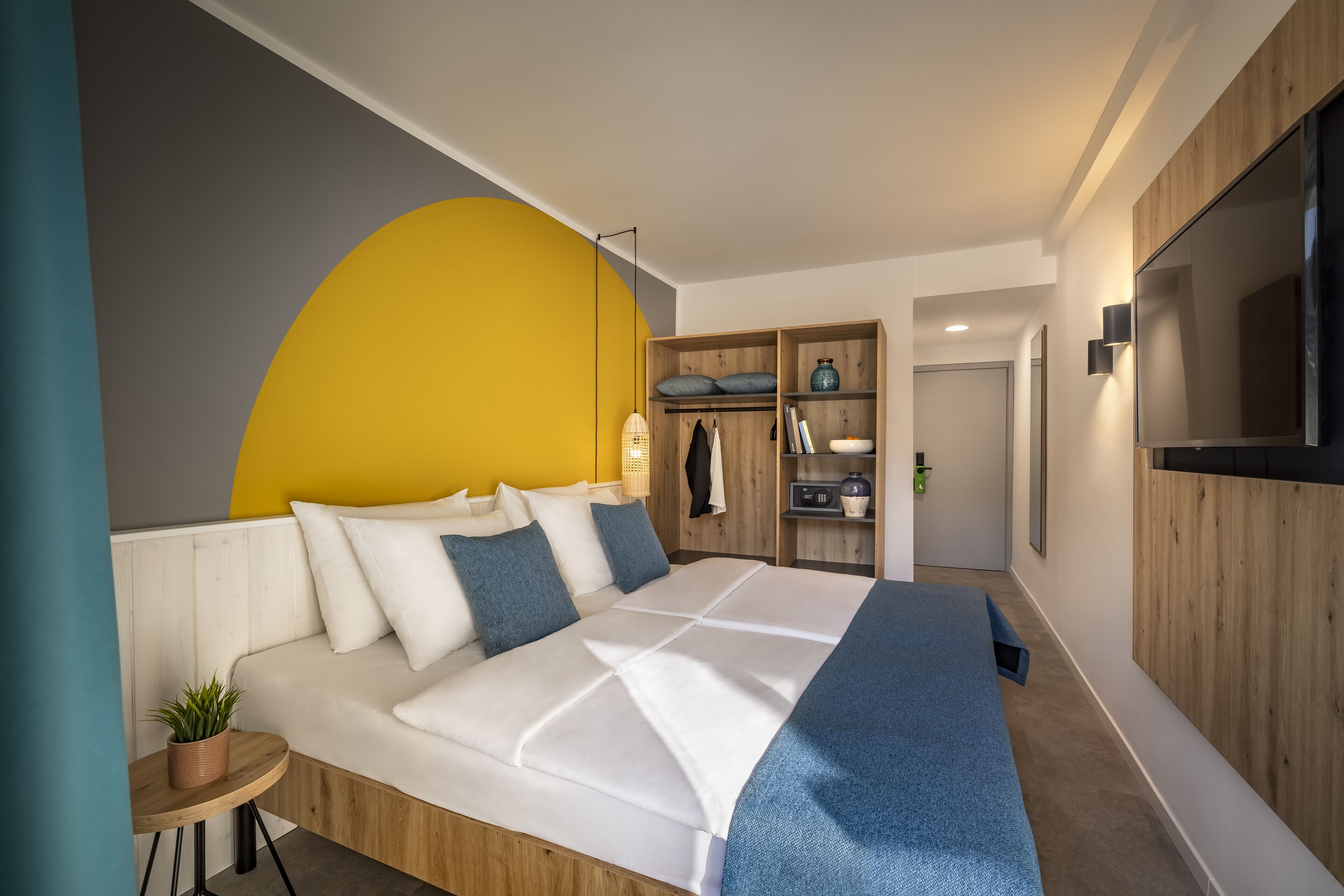 A modern bedroom with a large bed, a wooden wardrobe, a TV, and a yellow accent wall.
